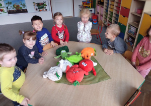 dzieci na zdjęciu stoją przy stoliku z pluszowymi warzywami