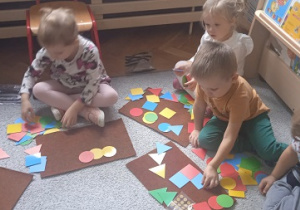 dzieci układają na chodniczkach papierowe figury geometryczne