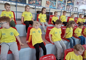 dzieci w żółtych koszulkach siedzą na trybunach basenu