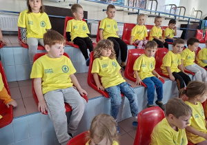 dzieci w żółtych koszulkach siedzą na trybunach basenu