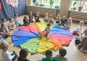 chłopiec siedzi na kolorowej chuście z torbą cukierków wokół niego siedzi grupa dzieci