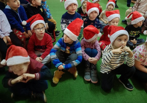 gromadka dzieci w czapkach mikołajkowych siedzi i słucha Mikołaja