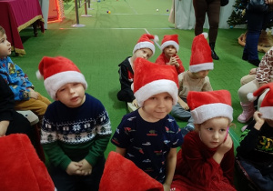 gromadka dzieci w czapkach mikołajkowych siedzi i słucha Mikołaja