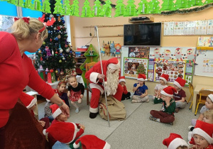 dzieci w mikołajkowych czapkach siedzą w półkolu i czekają na prezenty