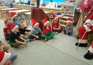 dzieci w mikołajkowych czapkach siedzą w półkolu i czekają na prezenty